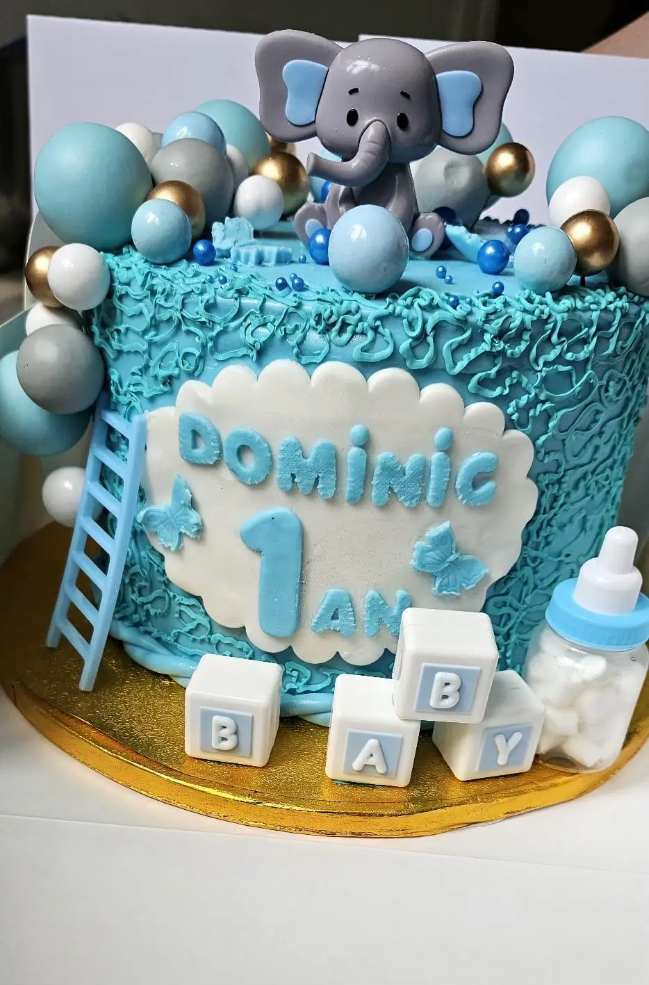 Blue baby celebration cake decorated with an elephant topper, balloons, baby blocks and bottle details
